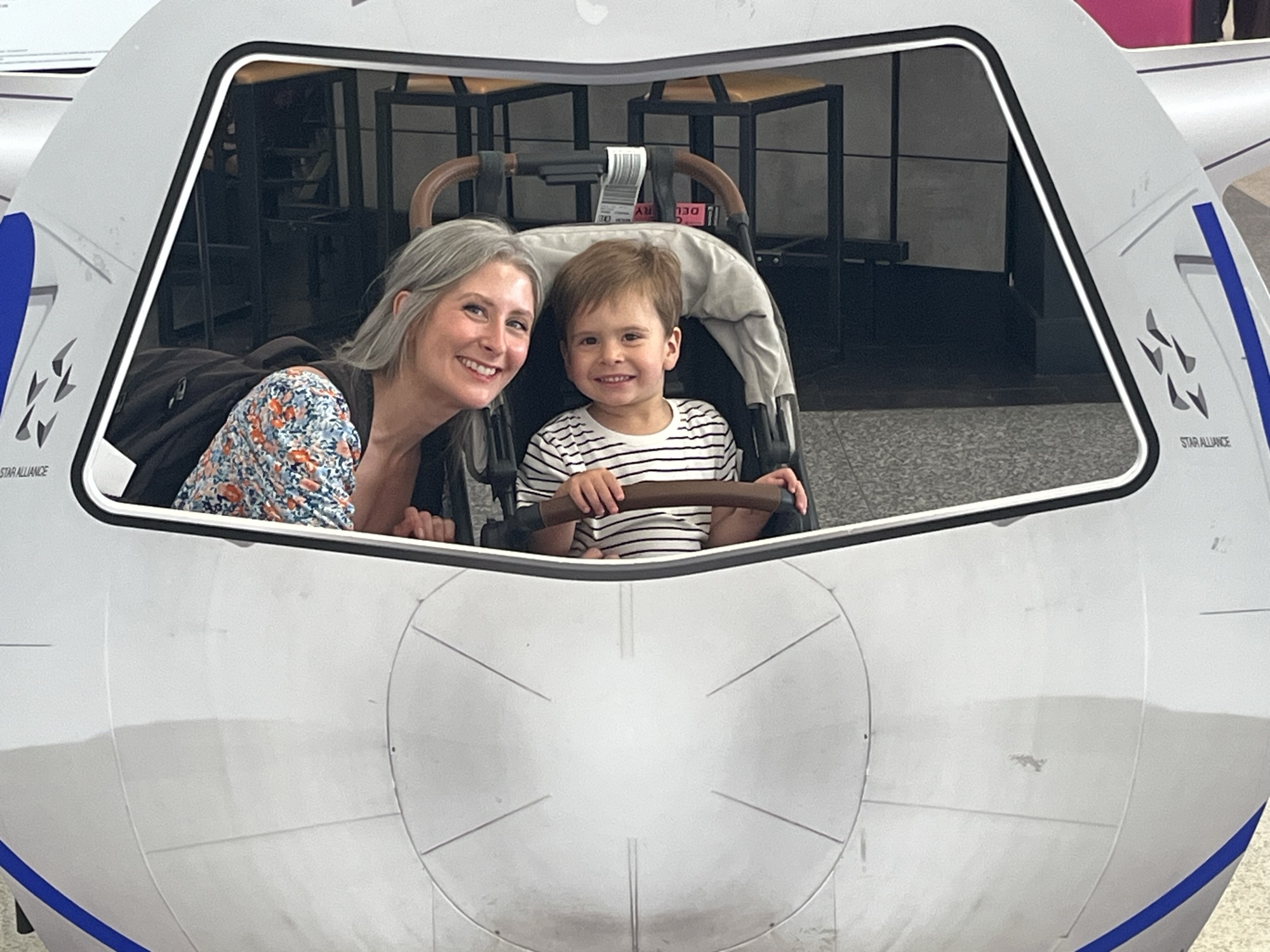 George at the wheel of a play airplane in the Newark airport terminal, grinning, with his mom leaning in beside him