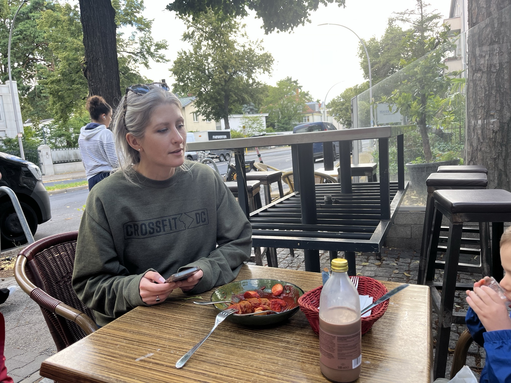 Liza at a sidewalk table with a bowl of currywurst and a bottle of chocolate milk, George sipping his at the edge of the frame
