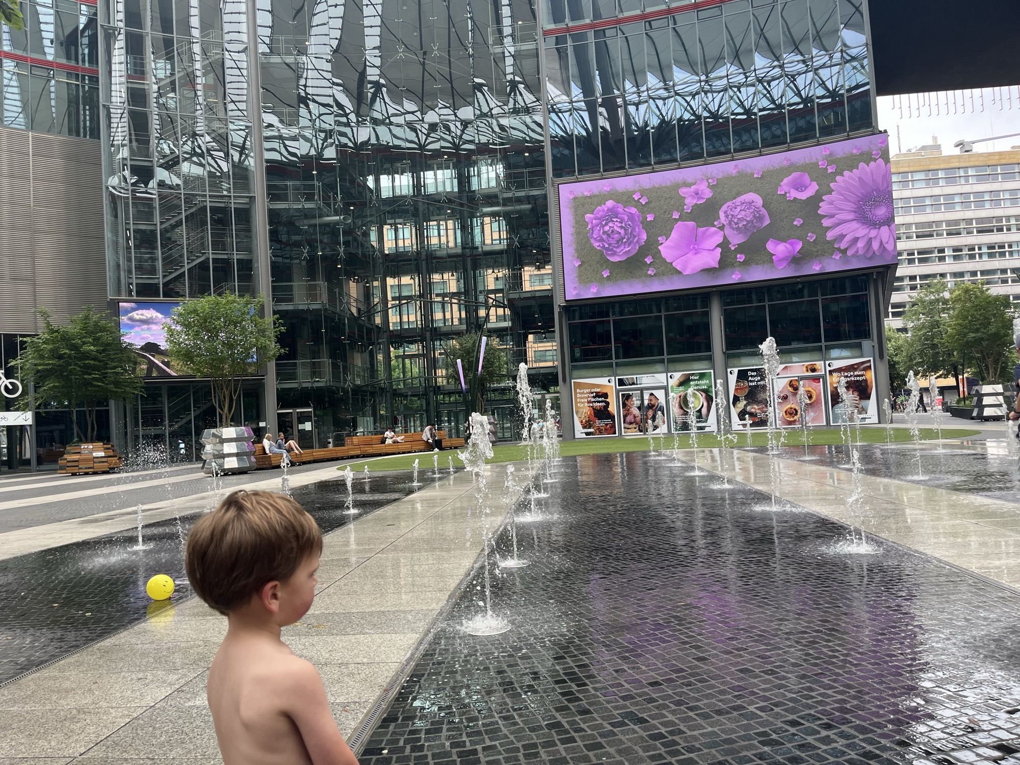 George at the edge of a splash fountain in a glass-walled plaza downtown, a yellow smiley-face ball floating in the water