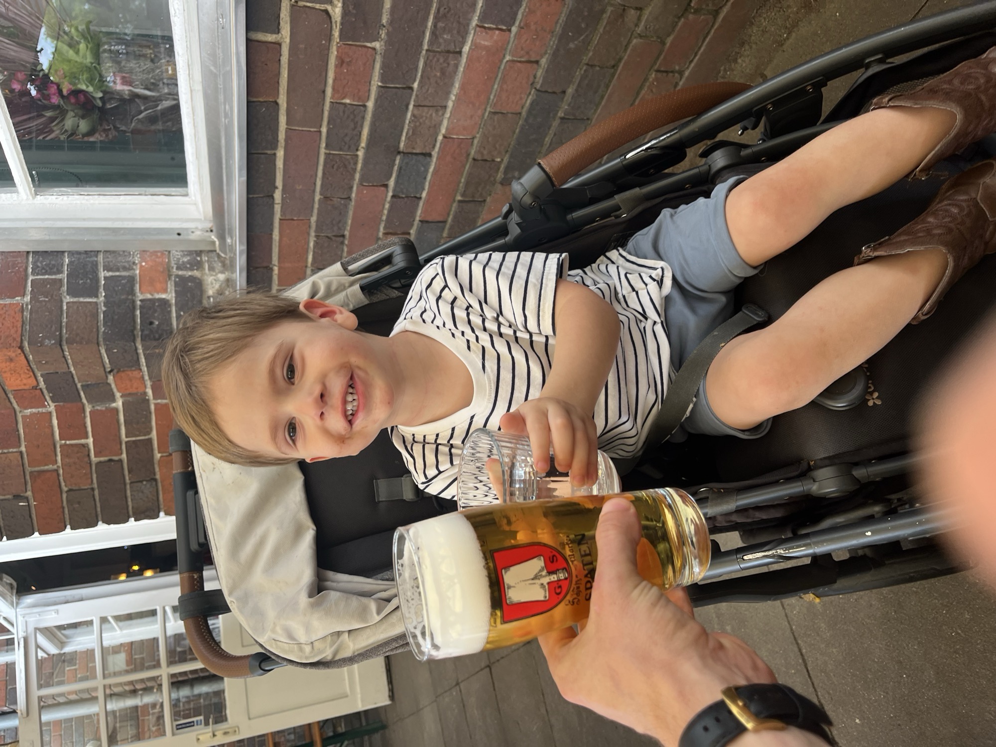 Georgie grinning in his stroller at Golgatha Biergarten, his sparkling water in hand beside my Spaten stein