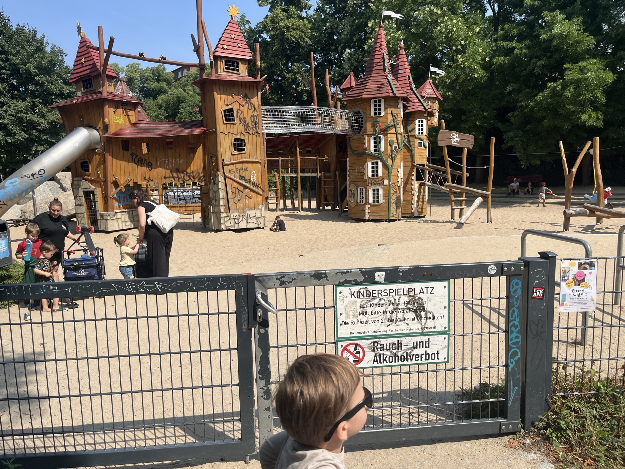 George at the gate of the fairytale playground in Heinrich-Lassen-Park, castle towers and slides rising behind the fence