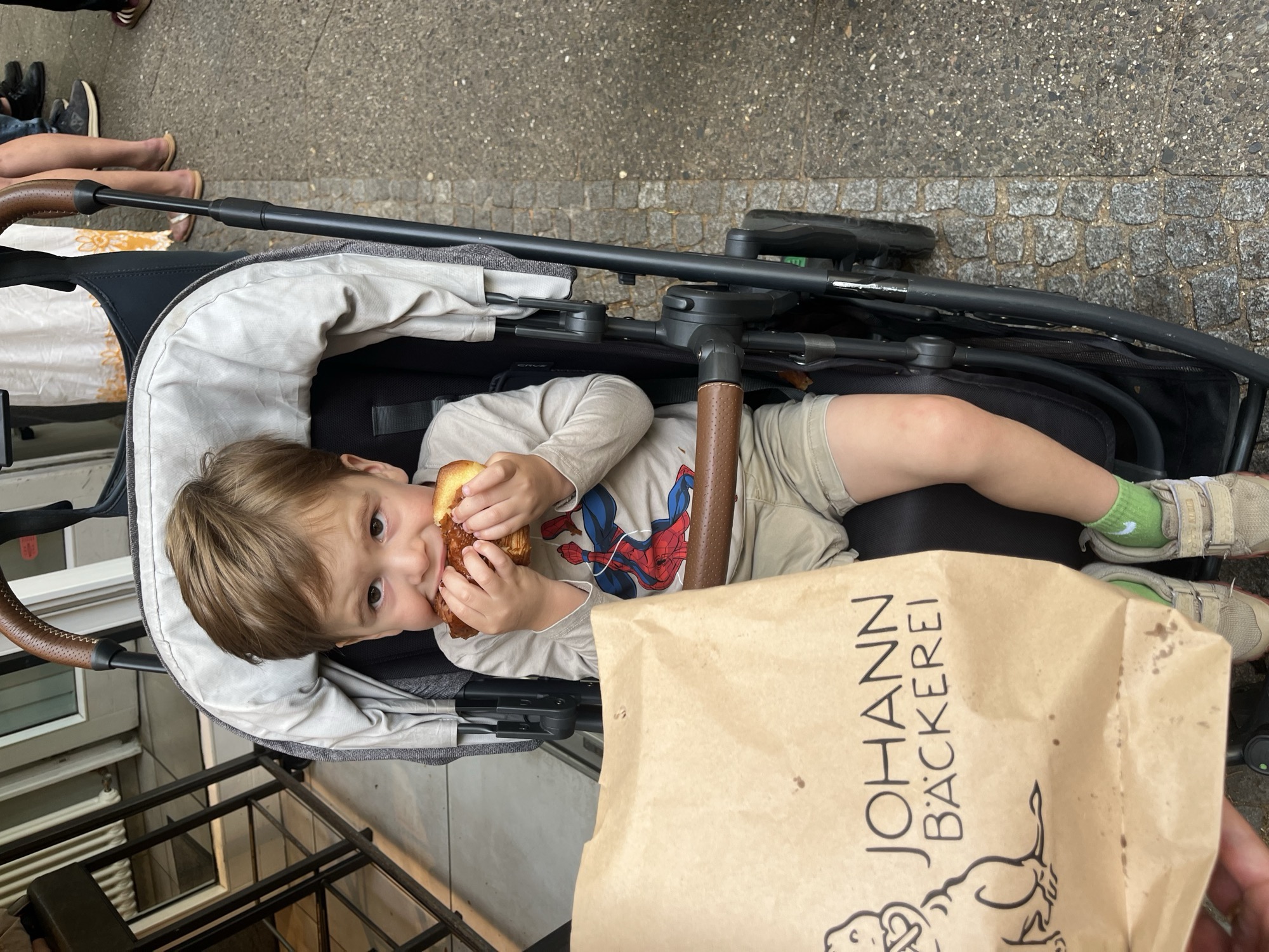 George eating a pain au chocolat in his stroller behind a Johann Bäckerei paper bag