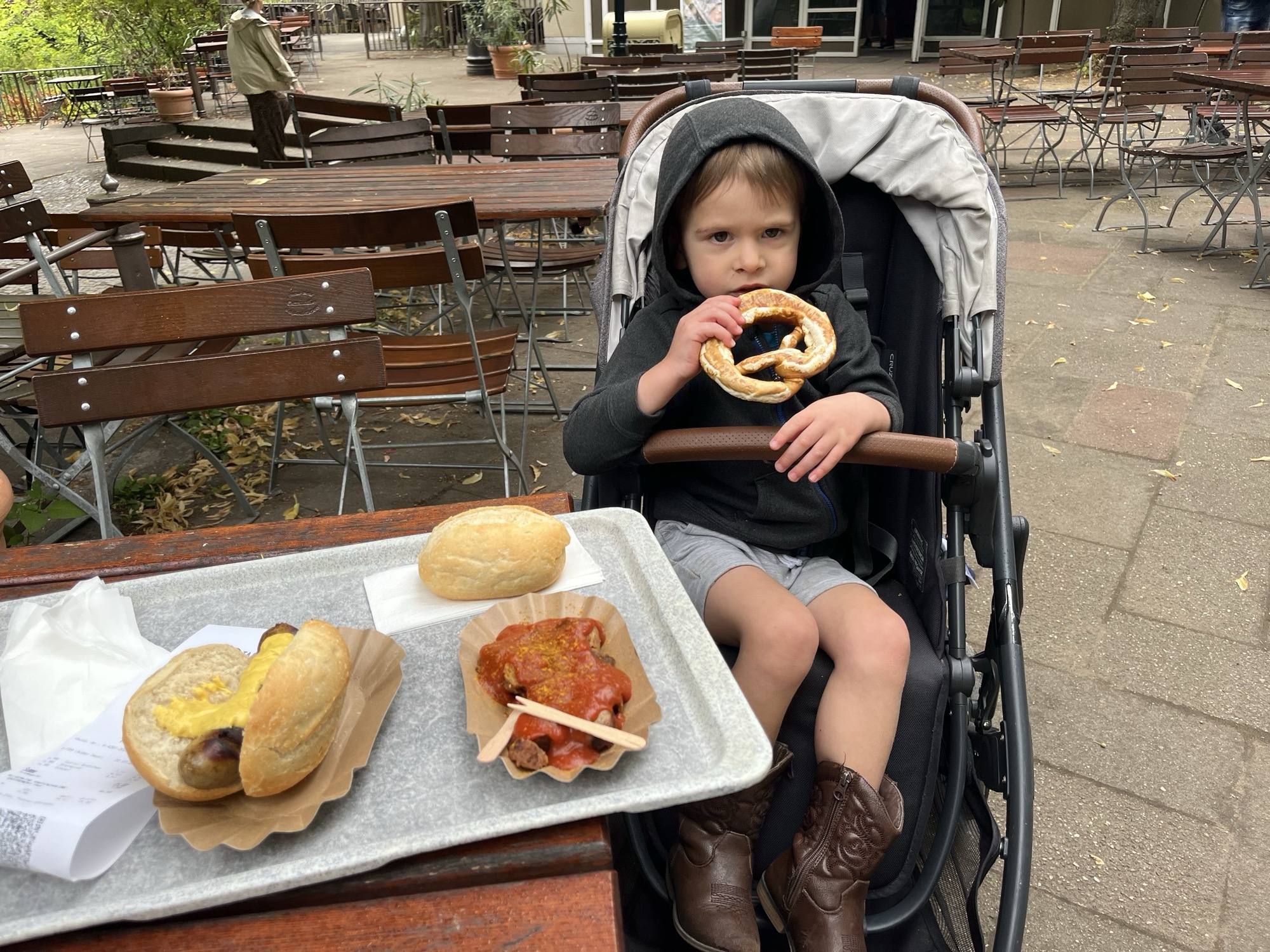 George in his stroller at the zoo, a pretzel in both hands, with a tray of bratwurst and currywurst on the table